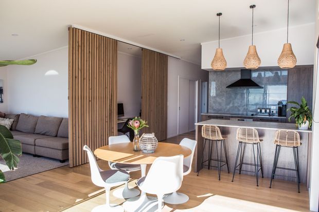 Open-plan contemporary kitchen, dining and living area with warm timber flooring, a wood dining table and white molded chairs in the foreground. A kitchen with a long island, dark cabinetry, reflective backsplash and woven bar stools occupies the right side, while vertical timber screening separates the living zone with a sectional sofa. Woven pendant lights and indoor plants add natural texture.
