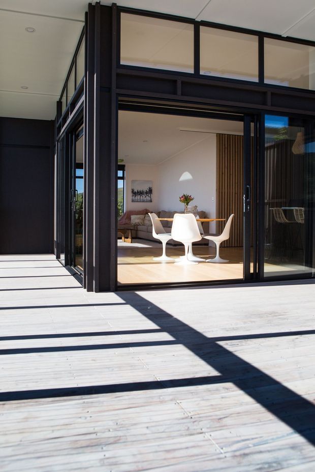 Exterior view of a contemporary home opening onto a broad timber deck. Black-framed floor-to-ceiling glazing and clerestory windows create a strong indoor-outdoor connection, with a bright living and dining area visible inside. The interior includes a white tulip-style dining set, sofa, timber slatted feature wall, and exposed timber flooring. Strong sunlight casts geometric shadows across the deck.