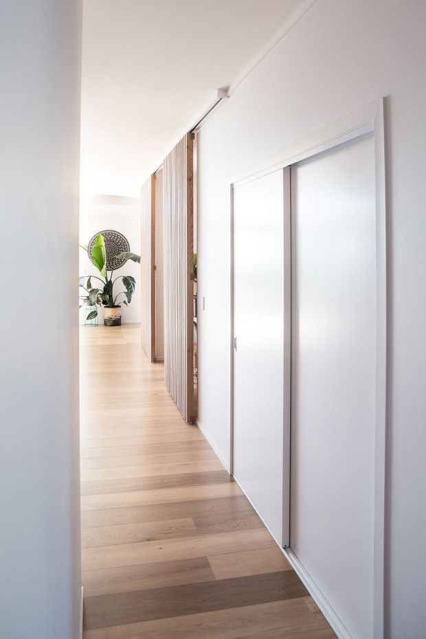A bright, narrow residential hallway with pale timber flooring, white walls and ceiling, and full-height white sliding doors along one side. Vertical timber screening and slatted partitions add texture, while a potted plant and decorative wall feature are visible at the far end.