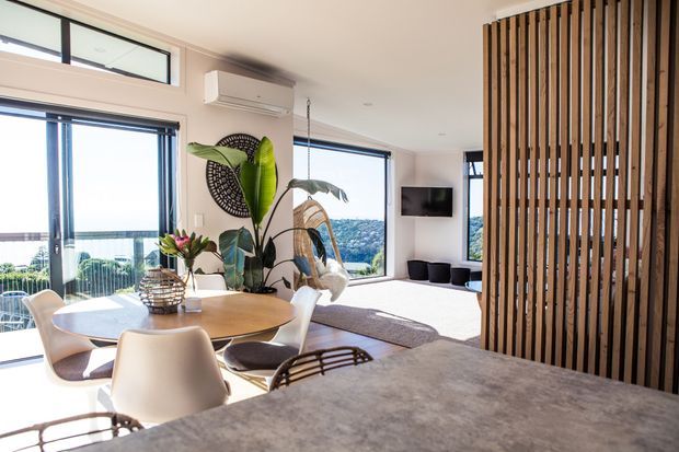 Bright open-plan dining and living area with floor-to-ceiling glazing, views toward a green hillside and water, a round timber dining table with white chairs, indoor tropical plants, a wall-mounted television, and a vertical timber slat screen dividing the spaces. A large grey stone-look benchtop or island occupies the foreground.