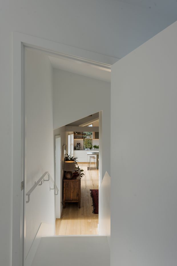 View from a white-painted stairwell or landing through an open doorway toward a bright kitchen and living area. The space features pale timber flooring, white walls and ceiling, a simple light-coloured stair handrail, timber cabinetry or console furniture, indoor plants, and a visible kitchen island in the distance. The composition emphasizes the vertical circulation space and interior sightline.