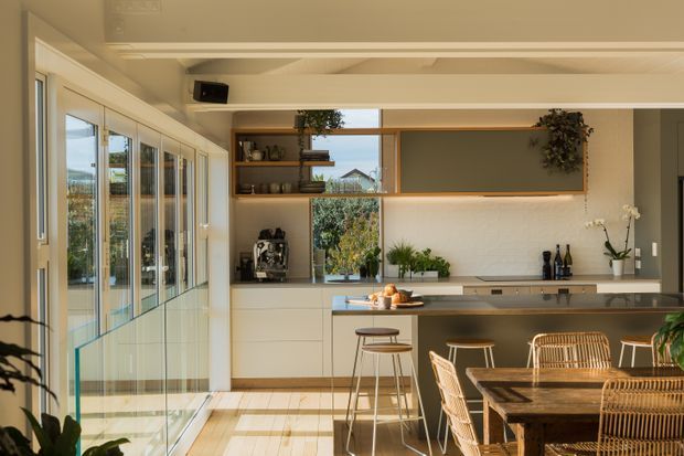 Warm, light-filled open-plan kitchen and dining area with extensive glazing along one side, pale timber flooring, white integrated cabinetry, a long island with stools, open timber shelving, and a built-in dining nook furnished with woven rattan chairs. Indoor plants and neutral finishes create a relaxed contemporary atmosphere.