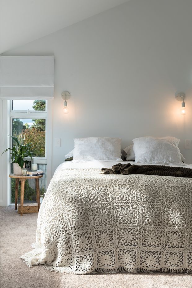 A bright, serene bedroom with a large bed dressed in white textured bedding and a crocheted floral-patterned bedspread. Pale grey walls, carpeted flooring, simple wall sconces, a window with a white Roman blind, and a small timber plant table create a calm, natural interior.