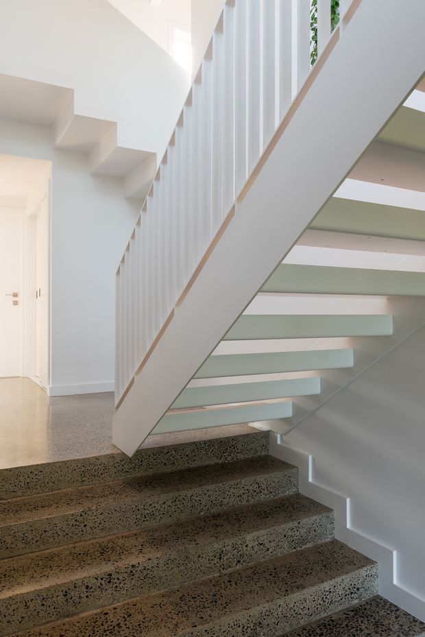 A bright contemporary stairway with broad terrazzo steps, a white stringer and balustrade, and translucent or pale glass stair elements. Vertical white screening forms a strong feature along the upper stair, while clean white walls and integrated skylight-like openings create an airy, minimalist interior.