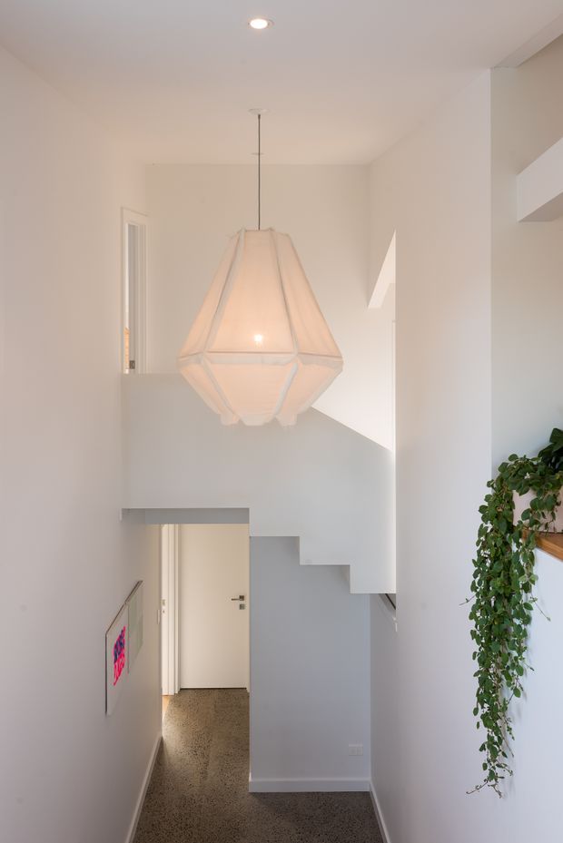 A bright, minimalist residential stair hall with white walls, a sculptural fabric pendant light, and a compact staircase descending to a lower hallway. A trailing indoor plant sits on a wall-mounted timber shelf, while a terrazzo-like dark floor and a colourful framed artwork add contrast.
