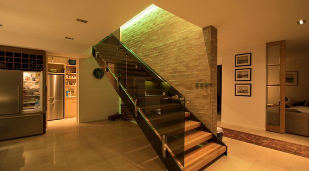 Warmly lit contemporary interior showing a dramatic open-riser staircase with dark timber treads, dark structural stringers and glass balustrades. A tall exposed brick feature wall rises beside the stairs to a skylit or clerestory-like opening. The space also includes a compact kitchen or refreshment area with stainless-steel appliances and built-in shelving, and a bedroom visible through a doorway on the right.