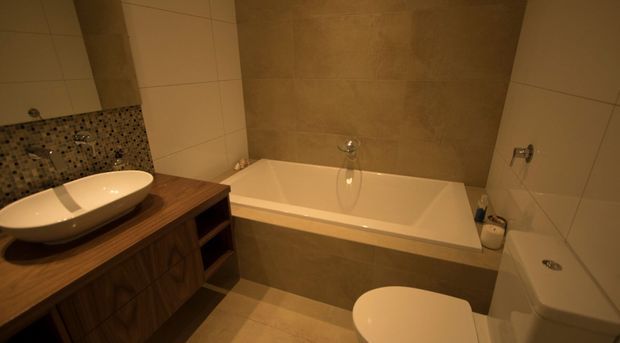 Warmly lit contemporary bathroom featuring a freestanding white vessel basin on a timber vanity, a built-in bathtub with a large stone-look feature wall, tiled walls, mosaic splashback, and a partially visible toilet and shower area.