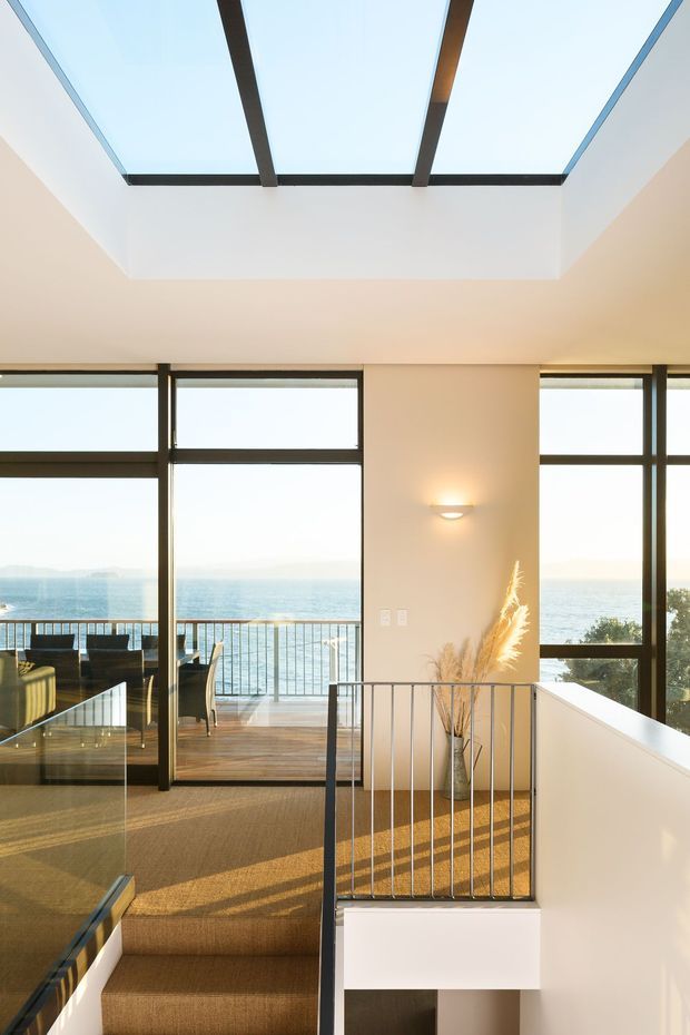 Bright contemporary interior landing or stairway overlooking a waterfront through expansive dark-framed glazing. A large skylight with exposed dark structural beams illuminates the double-height circulation space. Carpeted stairs, glass and metal balustrades, white walls, timber balcony decking, and a minimalist wall sconce are visible, with a small dining or living area beyond the glazing.