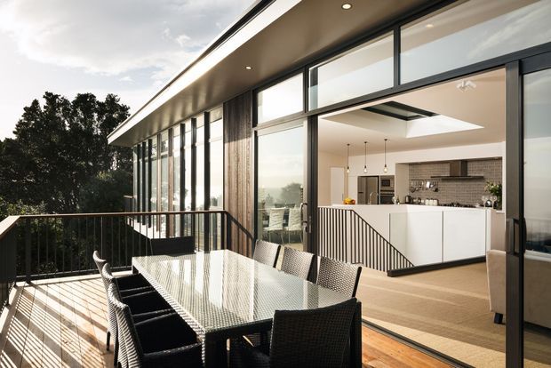 A contemporary open-plan kitchen and dining area opening onto a spacious outdoor terrace. The home features extensive floor-to-ceiling glazing, dark slender window and door frames, warm timber flooring, a white island with integrated cabinetry, a grey brick-pattern splashback, and a large skylight. Outdoor dining furniture and black metal balustrades occupy the terrace, with leafy vegetation beyond.
