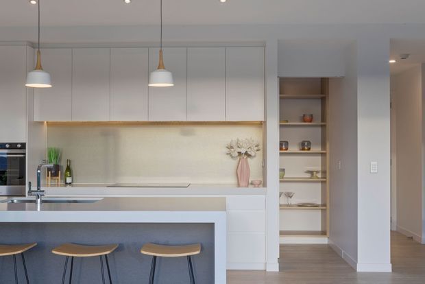 Bright contemporary kitchen with a central island and three stools, white handleless cabinetry, a pale textured splashback, integrated appliances, warm timber accents, pendant lights, and open display shelving. A hallway is visible to the right.
