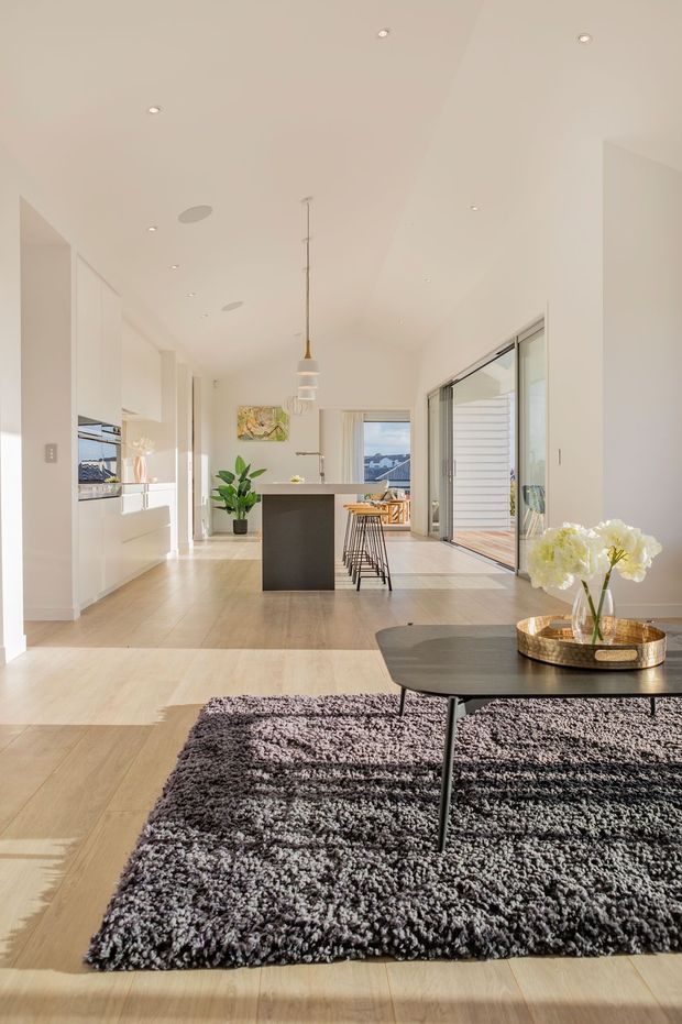 Bright open-plan contemporary interior combining a living area and kitchen. The space features white full-height cabinetry, a dark kitchen island with stools, pale timber flooring, expansive sliding glass doors, a black coffee table, and a textured charcoal rug. Natural light fills the room, with a view toward an outdoor balcony or terrace beyond the glazing.