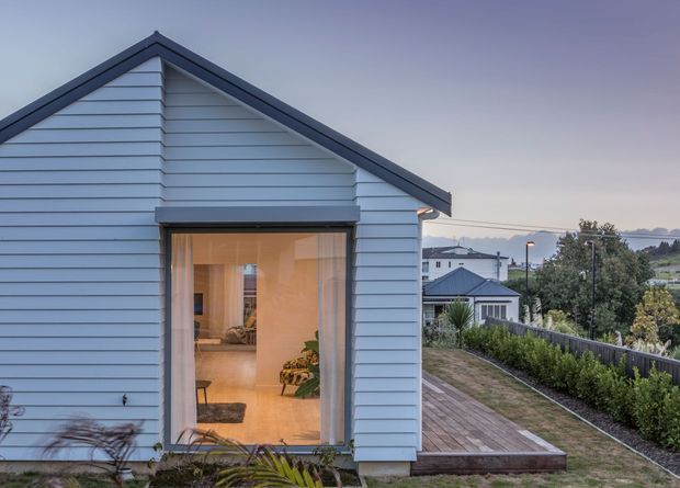 Exterior view of a small single-storey gable-roof structure clad in pale blue horizontal weatherboards. A large glazed opening reveals a warmly lit interior, while a timber deck extends along the side beside landscaped planting and a fenced suburban setting.