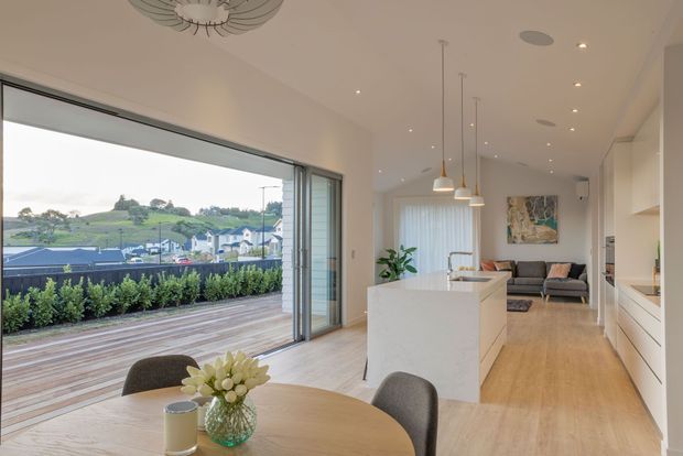 Bright open-plan interior combining a dining area, kitchen and living room, with pale timber flooring, white cabinetry and a large island. Full-height sliding glass doors open onto a timber deck overlooking a landscaped suburban setting. The room features pendant lighting, a neutral sofa, contemporary artwork and restrained decorative styling.