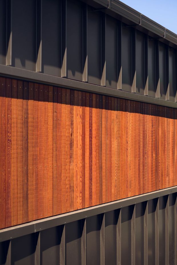 Close-up exterior detail of a contemporary building facade, combining warm vertical timber boarding with dark grey vertical profiled metal cladding. Strong sunlight creates pronounced shadows across the ribbed upper and lower cladding, with a dark metal roof edge visible at the top.