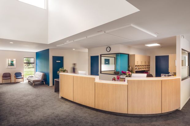 A bright contemporary reception area with a large curved timber reception desk, blue accent walls and doors, grey carpet, and a partially visible waiting area with lounge chairs. The space has a high ceiling with a large recessed or void-like opening, integrated linear lighting, clerestory-style glazing, and built-in shelving behind the desk.
