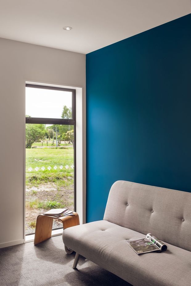A compact, contemporary interior corner with a deep blue feature wall, light neutral walls and ceiling, and a large vertical window overlooking greenery. A light grey upholstered button-tufted sofa occupies the foreground, accompanied by a small curved timber side table with magazines. Recessed ceiling lighting and neutral carpeting complete the restrained, modern setting.