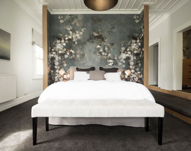 A refined bedroom centred on a large upholstered bed, with a dramatic floral feature wall framed by timber posts. The room has dark carpet, white bedding, layered neutral textiles, wall-mounted bedside lights, a coffered or panelled white ceiling, and built-in timber joinery visible to the right.