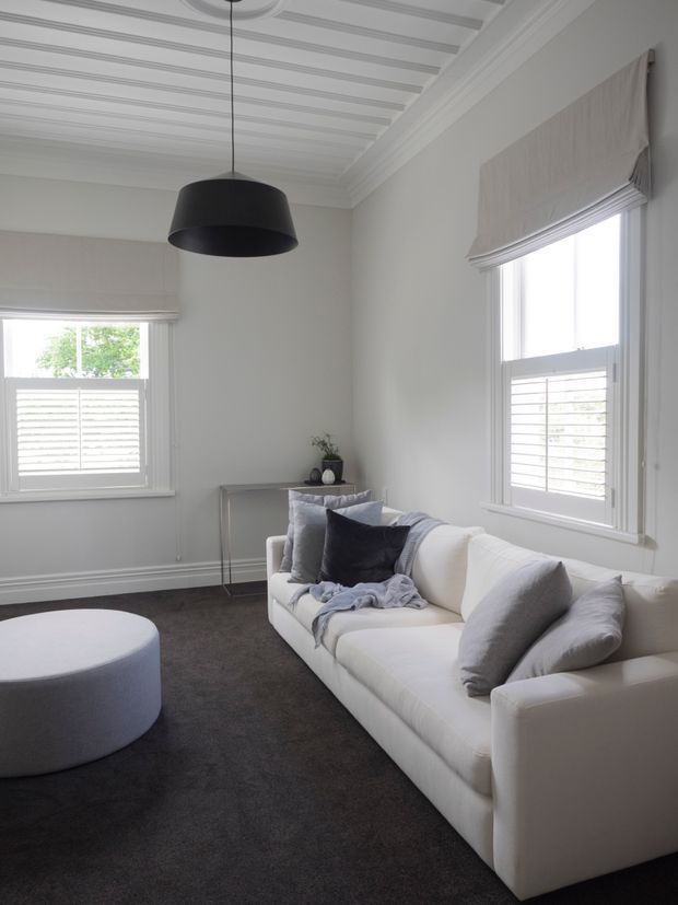 A bright, refined living room with a white upholstered sofa, grey cushions and throw, round upholstered ottoman, dark carpet, and a slim console table. The room features tall shuttered windows with fabric Roman blinds, white decorative wall mouldings, and an ornate coffered or panelled ceiling with a large black pendant light.