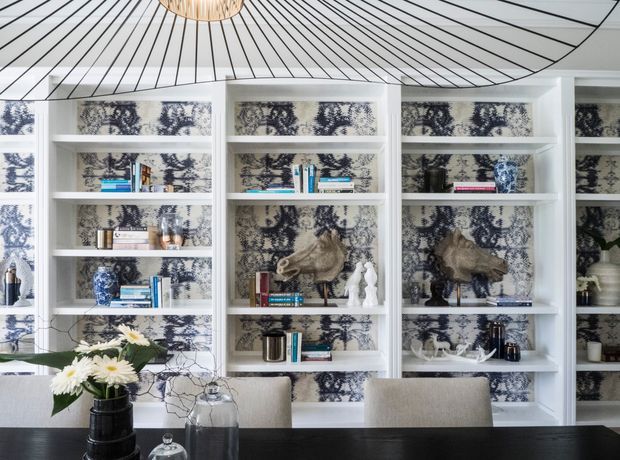 A refined dining area with a dark dining table and upholstered chairs positioned in front of extensive white built-in shelving. The shelving is backed with blue, grey, and cream patterned wallpaper and styled with books, ceramics, sculptures, vases, and decorative objects. A striking oversized pendant light with a radial ribbed shade hangs above, while a floral arrangement provides a natural accent.