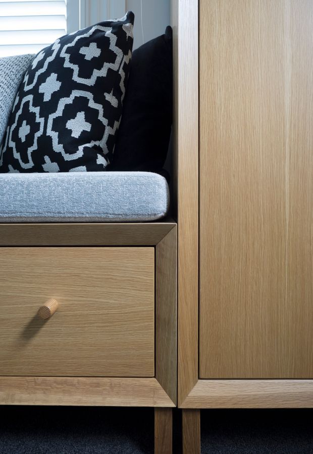 Close-up detail of custom timber joinery featuring a built-in upholstered bench seat with light grey cushion and black-and-white patterned cushions, above a timber drawer and beside a tall timber cabinet door. White plantation shutters are visible in the background, and dark carpet appears below.