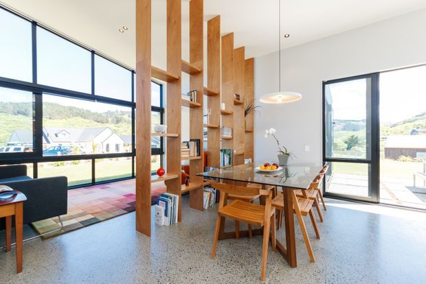 Bright contemporary dining area with a glass-top timber dining table and matching wooden chairs, opening onto a spacious glazed living zone and terrace. Full-height black-framed windows and doors provide expansive views, while a tall timber open shelving partition adds warmth and vertical definition. The room has a polished terrazzo or concrete-look floor, white walls and ceiling, and a simple pendant light.