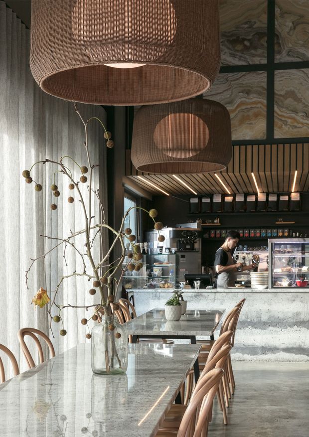 A refined contemporary café interior with a long stone communal dining table, curved timber chairs, a concrete service counter, dark shelving, and large woven pendant lights. Tall sheer curtains soften the daylight, while decorative branches and layered stone wall panels add warmth and texture. A barista counter and coffee equipment are visible at the rear.