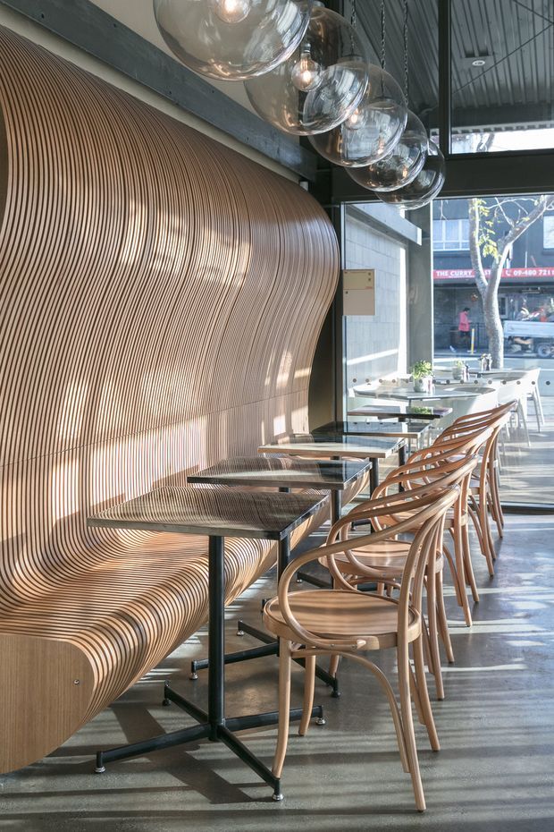 A narrow contemporary café or restaurant interior with a long sculptural ribbed timber banquette flowing into a curved wall feature. Small square tables with black metal bases line the banquette, paired with bentwood dining chairs. Large spherical glass pendant lights hang in a row overhead. Full-height glazing opens onto the street, while the exposed dark ceiling and structural elements add an industrial character.