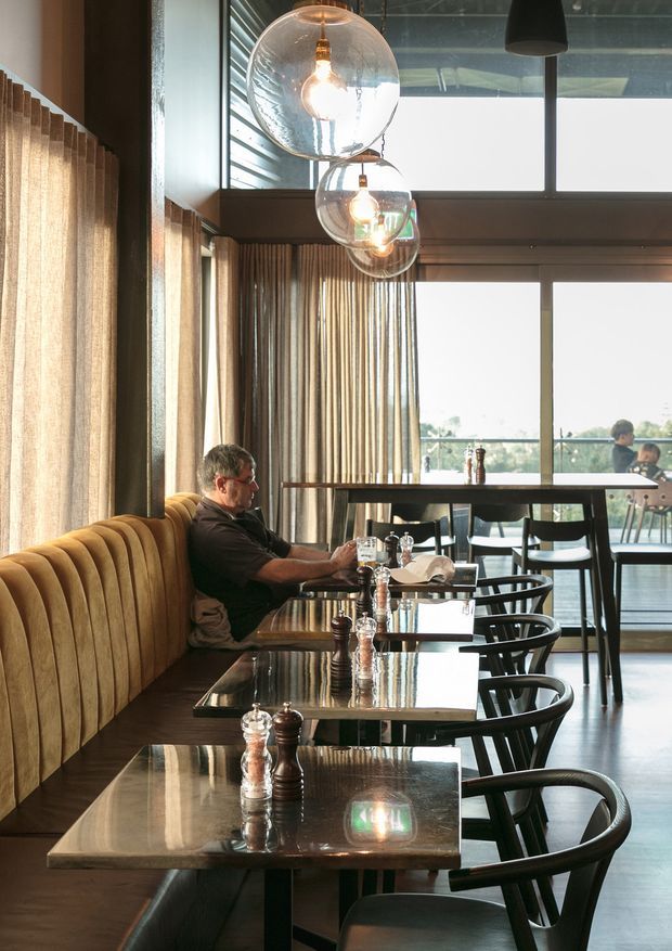 Interior view of a contemporary restaurant or café dining space with a long upholstered banquette, repeated dark dining tables and chairs, exposed pendant bulbs in glass globes, tall windows, and vertical curtains. The space has a warm, refined atmosphere with strong natural light and views to an outdoor balcony or terrace.