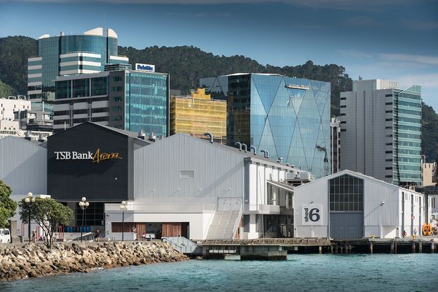 A waterfront urban commercial precinct with a large corrugated-metal event or exhibition building in the foreground, marked with a prominent venue number 6 and TSB Bank Arena signage. Modern multi-storey office buildings, a glass-facade tower, and a forested hillside form the background, while rocky shoreline and calm water occupy the lower edge.