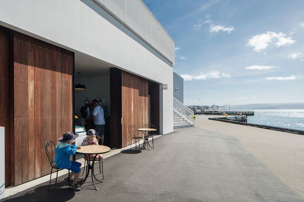 A contemporary waterfront commercial or hospitality building with a stark white and grey exterior, large dark timber sliding or folding doors, and an expansive paved terrace. Outdoor café tables and chairs sit beside the open doors, with a small harbour, pier, and water visible beyond. The image is brightly lit with a clean, relaxed coastal atmosphere.