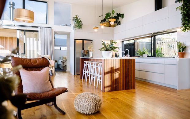 Bright open-plan kitchen and living area with warm timber flooring, white cabinetry, a timber island with bar stools, leather lounge seating, indoor plants, pendant lighting, and large windows. The kitchen opens directly into a high-ceilinged living space with a clean contemporary design and warm natural finishes.