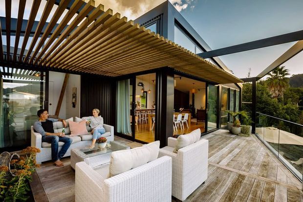 A contemporary multi-level home with a spacious outdoor terrace overlooking a wooded landscape. The terrace features white woven lounge seating, a glass balustrade, timber-look decking, potted plants, and a dramatic timber pergola with closely spaced slats. Open glazed doors connect the terrace to a warm interior dining and living area, while dark-framed glazing and projecting roof forms emphasize the modern architectural design.