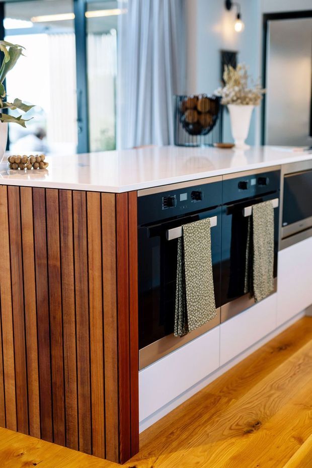 Close-up view of a contemporary kitchen island with a bright white benchtop, dark integrated ovens, white lower cabinetry, and a warm vertical timber slat end panel. Honey-toned timber flooring, reflective glazing, sheer curtains, and minimal decorative styling are visible in the background.