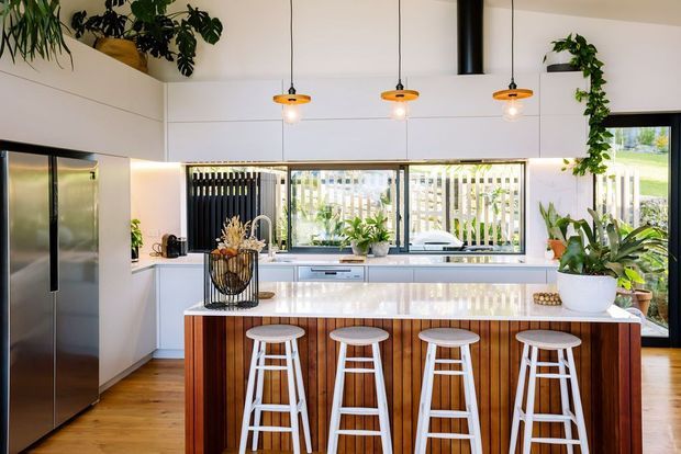 Bright contemporary kitchen with a large white island featuring vertical timber cladding, four white stools, white cabinetry, stainless-steel double refrigerator, timber flooring, pendant lighting, indoor plants, and wide windows overlooking an outdoor area.