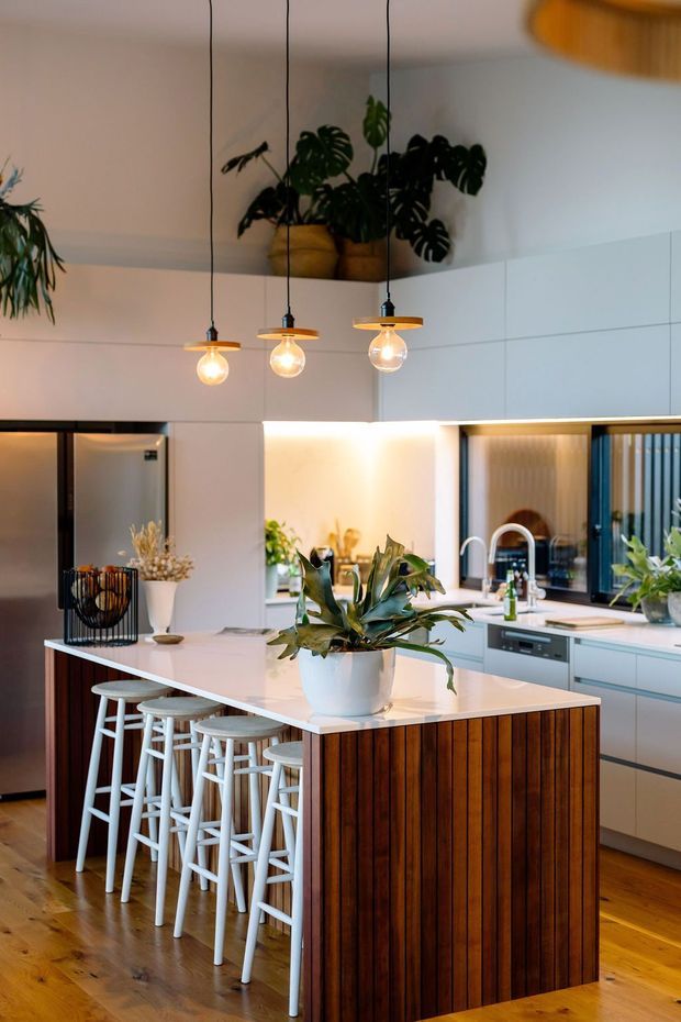 A bright, contemporary kitchen featuring a large white island with a vertically slatted dark timber base, white bar stools, integrated cabinetry, stainless-steel appliances, and a pale benchtop. Three pendant lights hang above the island, while indoor plants add a natural accent. Timber flooring and warm under-cabinet lighting create an inviting atmosphere.