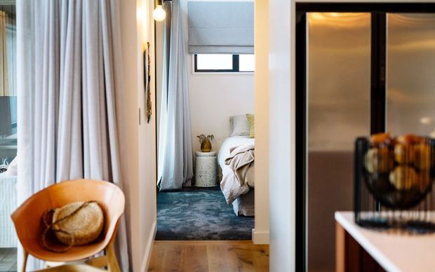 Interior view through a doorway into a compact bedroom, with floor-to-ceiling pale curtains, a warm timber floor, a blue patterned rug, a partially visible bed, and a sculptural orange armchair in the foreground. A dark-framed glazed or mirrored opening and console appear on the right, adding contrast to the otherwise light, softly styled space.