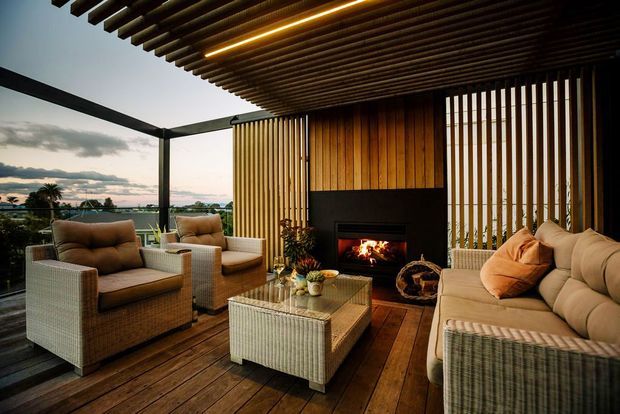 A covered outdoor lounge terrace with timber decking, a slatted timber pergola ceiling, vertical timber screening, and a built-in fireplace. Wicker armchairs and a large sectional sofa surround a glass-topped coffee table, with an open elevated outlook beyond the terrace.