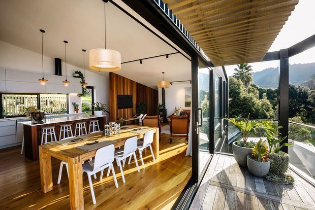 Bright open-plan contemporary dining and kitchen interior opening onto a covered timber deck or terrace. The room features warm timber flooring and dining table, white dining chairs, a kitchen island with bar seating, pendant lighting, black-framed full-height glazing, and a vertical timber feature wall. Large glass doors connect the interior to a planted outdoor terrace with views of a lush, hilly landscape.