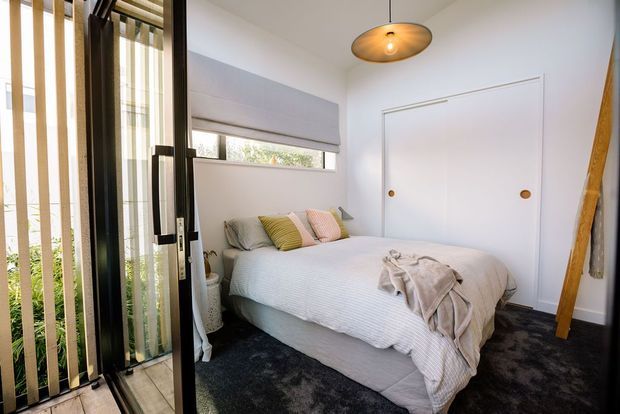 Compact, light-filled bedroom with a low-profile bed, soft neutral bedding, blush and mustard accent cushions, dark carpet, white walls, a narrow horizontal window with a Roman blind, and concealed built-in wardrobe doors. A black-framed glazed door with vertical blinds opens to the exterior, while a sculptural pendant light and leaning timber element add warmth.