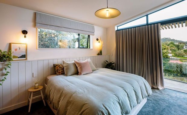 A warm, contemporary bedroom with a large upholstered bed dressed in pale blue bedding, soft neutral cushions, and full-height grey curtains. White vertical wall panelling, integrated bedside joinery, timber accents, pendant lighting, and expansive corner glazing create a bright, comfortable room with leafy views.