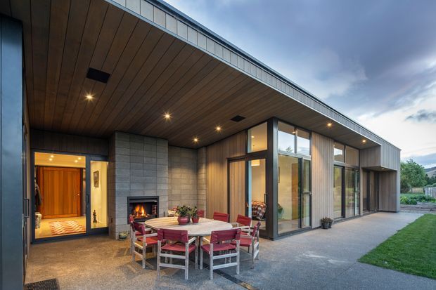 Contemporary single-storey home exterior with a deep sheltered patio and integrated outdoor dining area. The facade combines vertical timber cladding, grey masonry block walls, extensive dark-framed glazing, and a broad low-pitched roof with a timber-lined soffit. A built-in outdoor fireplace anchors the patio beside a large dining table, while the interior is visible through the open entry and glazing.