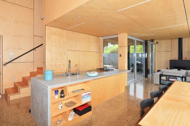 Open-plan contemporary kitchen and dining area with extensive light timber wall and ceiling panelling, a concrete island bench with open timber shelving, integrated stainless-steel sink and appliances, polished terrazzo-style flooring, and a compact staircase leading to an upper level. Full-height glazing connects the living area to the outdoors, while a dining table and seating are visible in the foreground.