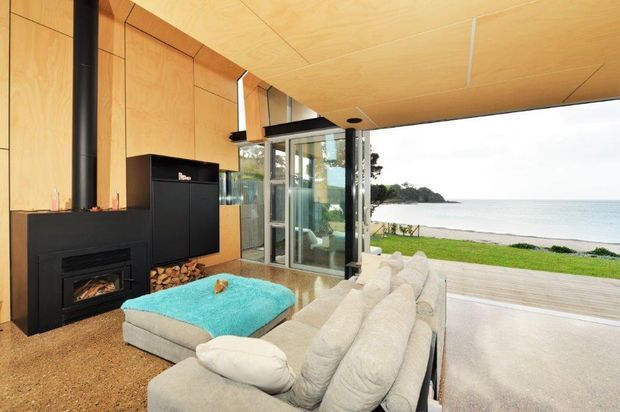 A warm, timber-lined living room opens directly onto a deck overlooking a beach and calm water. The room features a large light-colored sectional sofa, black wood-burning fireplace with exposed firewood storage, expansive glazed doors, and a turquoise ottoman. The ceiling and walls are clad in light plywood, creating a natural contemporary coastal interior with strong indoor-outdoor connection.
