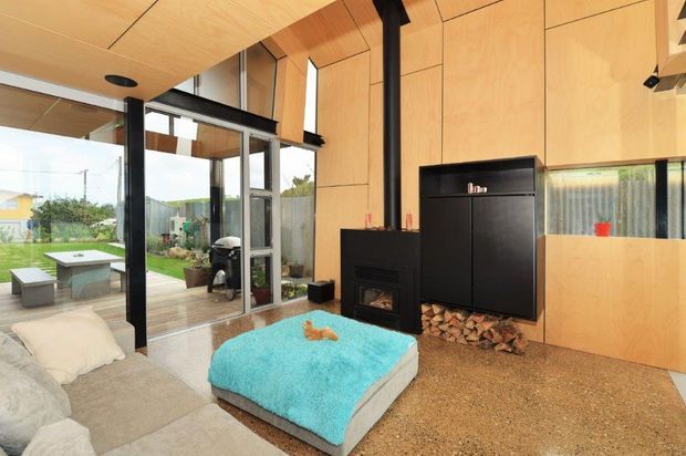 A contemporary open-plan living room with warm plywood wall and ceiling lining, black-framed floor-to-ceiling glazing, and a central black wood-burning fireplace with integrated firewood storage. The room opens onto a timber deck and landscaped garden, with a large neutral sectional sofa and bright turquoise ottoman in the foreground.