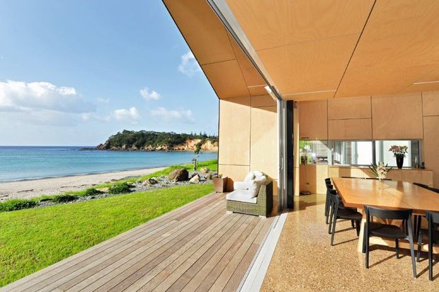 A bright contemporary open-plan dining and living space opening directly onto a timber deck beside a sandy beach and calm turquoise water. Full-height sliding glass doors create a seamless indoor-outdoor connection, while warm timber wall and ceiling panelling, timber cabinetry, and a natural-toned floor define the interior.