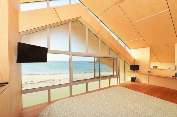 A bright, contemporary bedroom with a dramatic sloping timber-lined ceiling and extensive floor-to-ceiling glazing overlooking a sandy beach and ocean. The room includes a large bed, wall-mounted televisions, timber flooring, and streamlined built-in joinery.