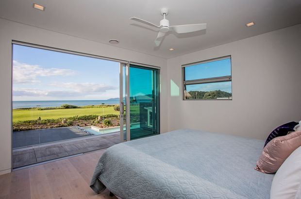 Bright, minimalist bedroom with a large bed, pale timber flooring, white walls and ceiling, a ceiling fan, recessed lighting, and extensive glazing. Sliding glass doors open to a paved terrace and landscaped grounds with a visible coastal or waterfront outlook.