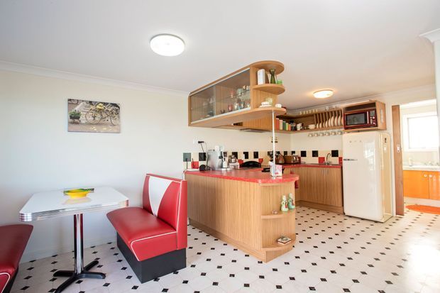 A bright retro-style kitchen and dining area with a compact built-in kitchen, curved timber breakfast peninsula, glass-front overhead cabinetry, red benchtop accents, checkerboard floor tiles, and a small white dining table with red upholstered chairs. The space includes a freestanding white refrigerator, microwave, coffee appliances, and a black-white-red geometric splashback.