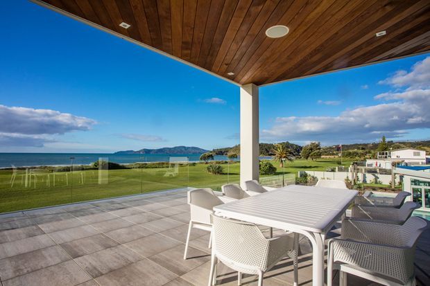 A spacious covered outdoor terrace overlooking a manicured lawn, swimming pool area, coastline, and distant hills. The terrace features grey tiled paving, a timber-lined ceiling with recessed lighting, frameless glass balustrades, and a white outdoor dining table surrounded by woven chairs. The setting is bright and coastal with expansive sea views.