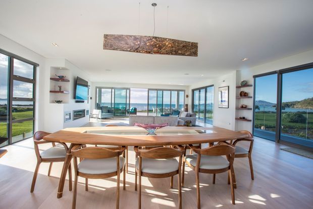 Bright, open-plan dining and living area with a long timber dining table and matching upholstered chairs in the foreground. Floor-to-ceiling glazing surrounds the room, providing expansive views toward water and landscaped grounds. The room has white walls and ceiling, pale timber flooring, built-in shelving, a contemporary pendant light, and a comfortable lounge area beyond the dining zone.